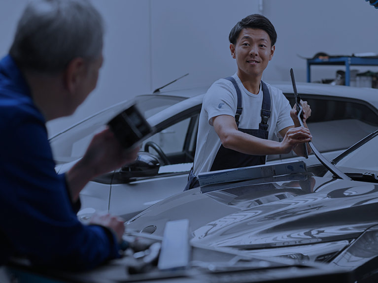 Two mechanics on a closed bonnet, one of whom is fitting a wiper blade